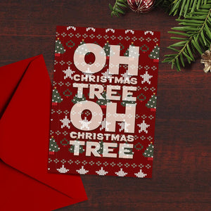 Card with Oh Christmas Tree written on it with a Christmas jumper background to the card. The card is next to a red envelope on a dark wood background with fir tree branches in shot.
